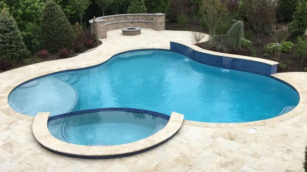 A backyard features a clean, curvy swimming pool with an attached circular hot tub. The pool, ideal for pool videos, is surrounded by tan stone tiles, with landscaping and a stone fire pit area in the background, inground swimming pools.