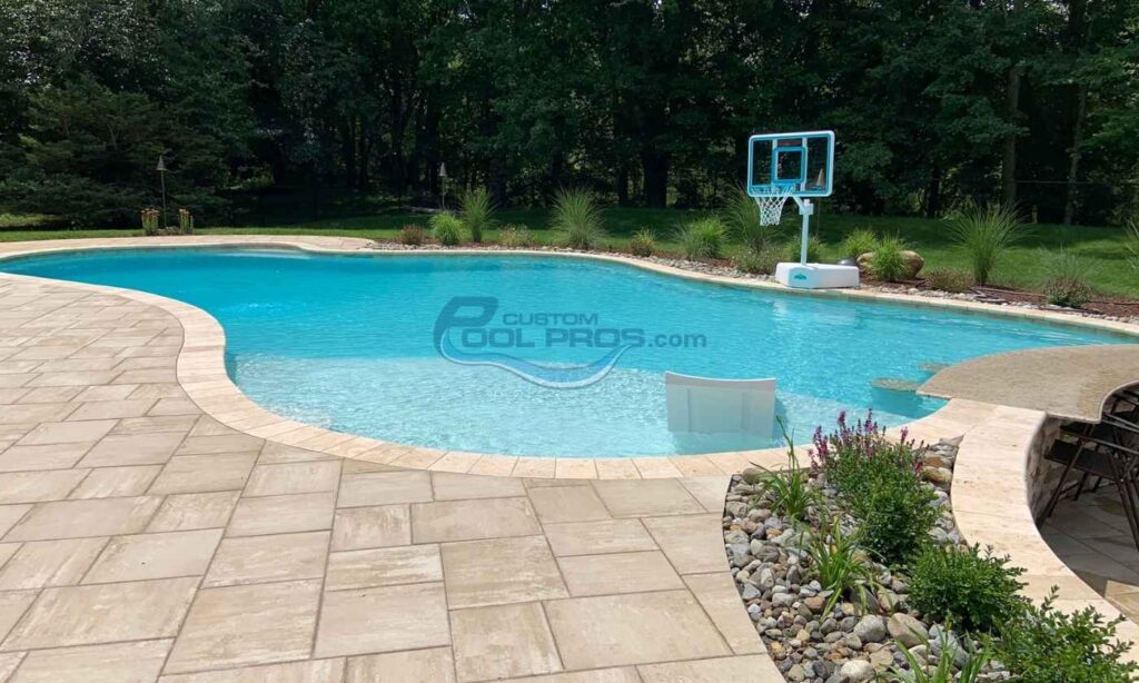 Backyard swimming pool with clear blue water, surrounded by a stone patio and landscaped greenery, featuring a basketball hoop for recreational activities. Pool contractors