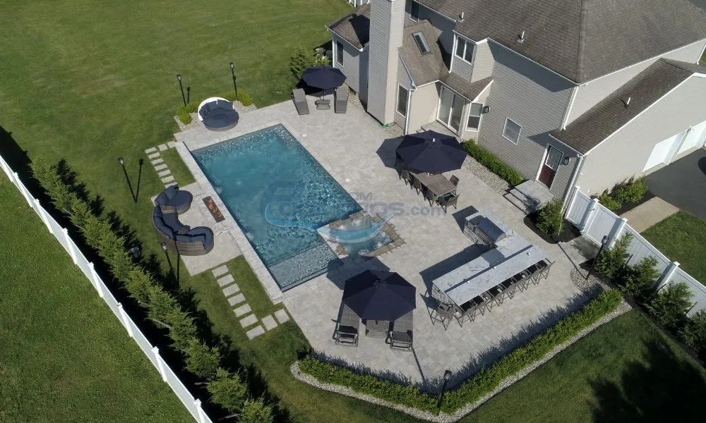 Aerial view of a modern backyard featuring a rectangular swimming pool surrounded by a stone patio, outdoor furniture including dining tables and lounge chairs, and a well-maintained lawn. The house in the background adds to the inviting outdoor setting.