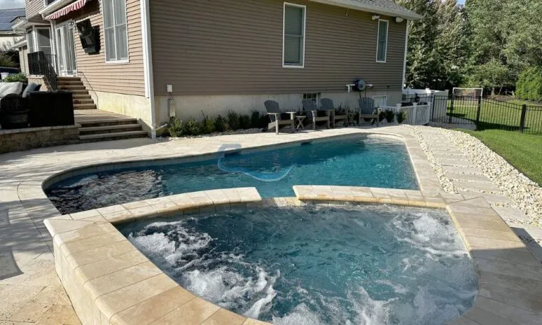 Backyard swimming pool with a spa, featuring a stone deck, surrounded by chairs and a house in the background. Ideal for relaxation and outdoor enjoyment, luxury pools