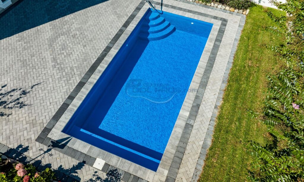 Aerial view of a modern rectangular swimming pool with clear blue water, surrounded by gray stone paving and a well-maintained green lawn. Ideal for outdoor relaxation and recreation, luxury pool builder.