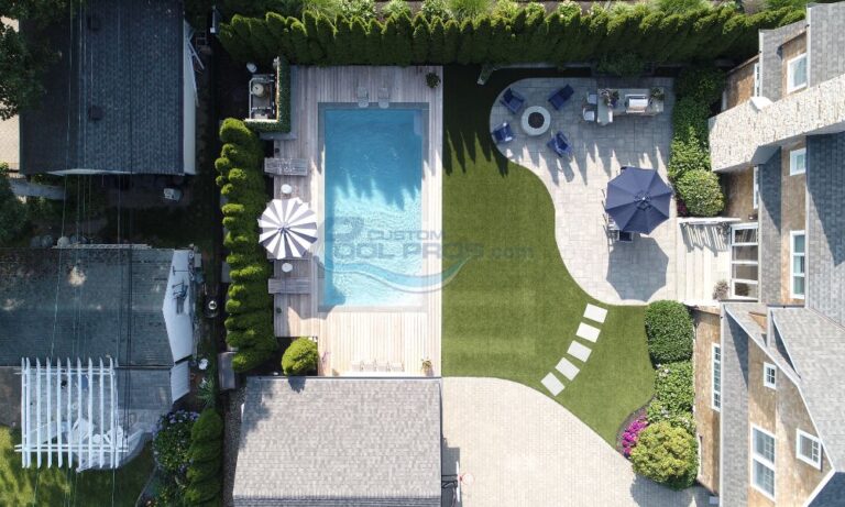 Aerial view of a modern backyard featuring a rectangular swimming pool surrounded by wooden decking, landscaped grass areas, and stone patios. The space includes lounge chairs, an umbrella, and outdoor seating for relaxation. Lush greenery and vibrant flowers enhance the outdoor aesthetic, creating a serene environment for leisure and entertainment. Backyard pools