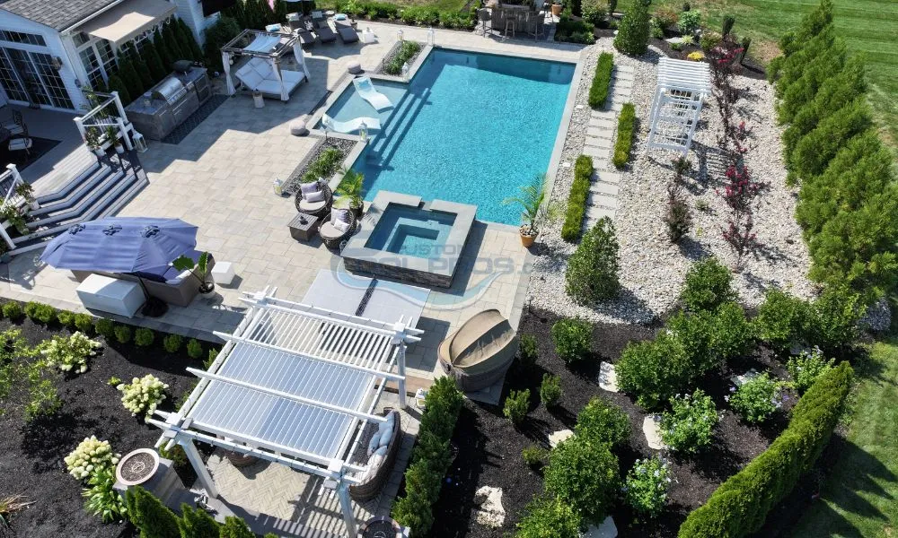 Aerial view of a beautifully landscaped backyard featuring a swimming pool, patio area with outdoor furniture, a pergola, and lush greenery. The design includes a hot tub, stone pathways, and decorative flowers, creating an inviting outdoor space perfect for relaxation and entertainment, pool contractor