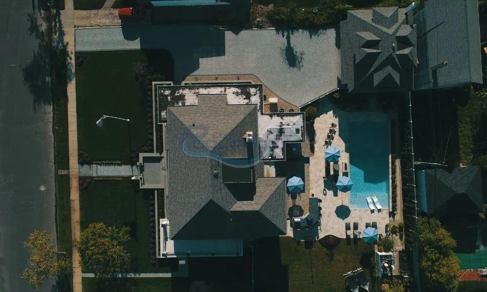 Aerial view of a modern residential property featuring a large swimming pool, sun loungers, and patio umbrellas, surrounded by landscaped gardens and driveways, pool contractors nj, backyard pool