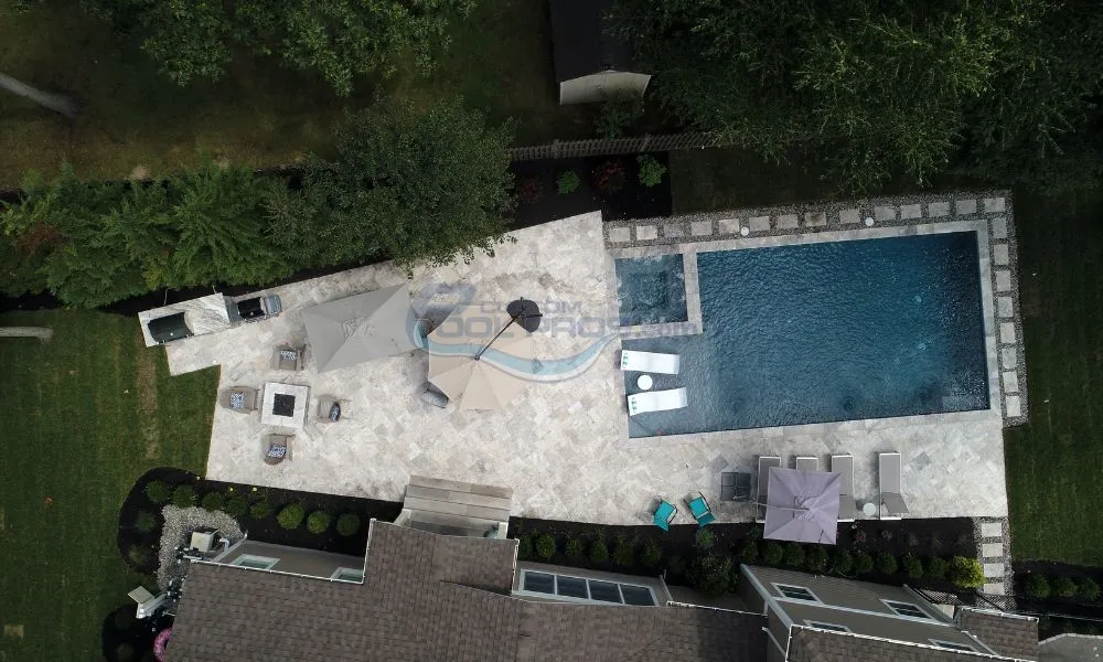 Aerial view of a modern backyard featuring a swimming pool, patio area with lounge chairs, and shaded seating under umbrellas, surrounded by lush greenery. Custom pool builders.