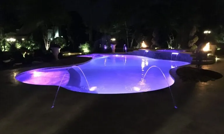 Illuminated outdoor swimming pool at night, featuring vibrant purple lighting and water fountains, surrounded by lush greenery, swimming pool installation new jersey