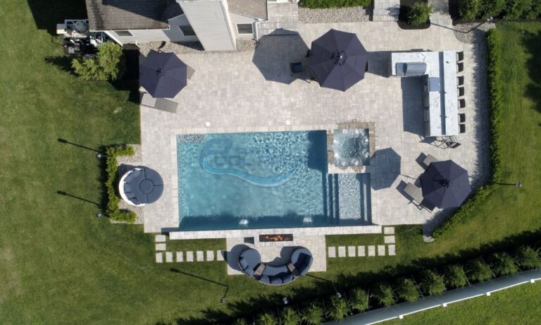 Aerial view of a luxurious backyard featuring a rectangular swimming pool, hot tub, and several dark umbrellas over lounge chairs, surrounded by well-maintained grass and stone pathways, forked river