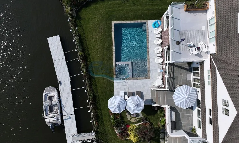 Aerial view of a luxurious backyard featuring a swimming pool, lounge chairs, and umbrellas, adjacent to a docked boat along a serene waterway, inground swimming pool, inground pool builders, pool builders nj